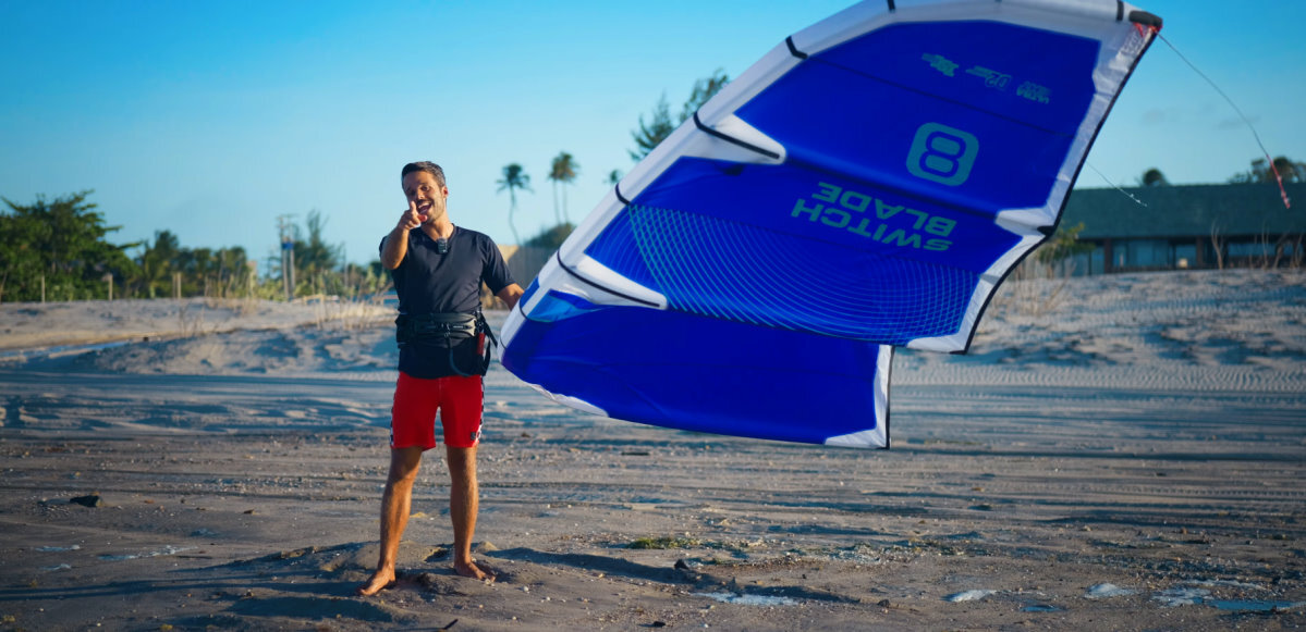 Rubens points at the camera as he holds a 2026 Cabrinha Switchblade kite in one hand.