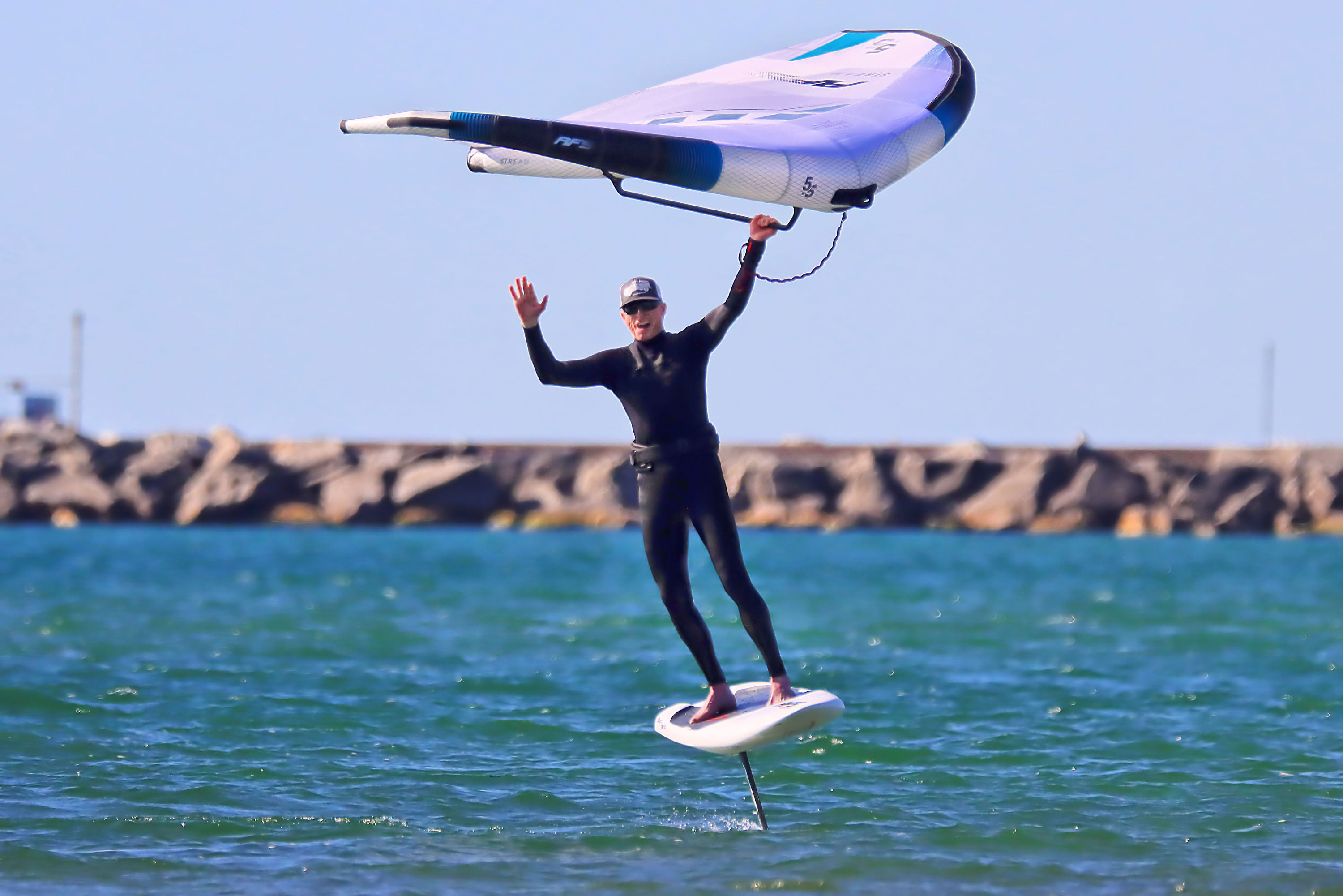 Jeff waves and smiles as he wingfoils, holding the wing over his head.