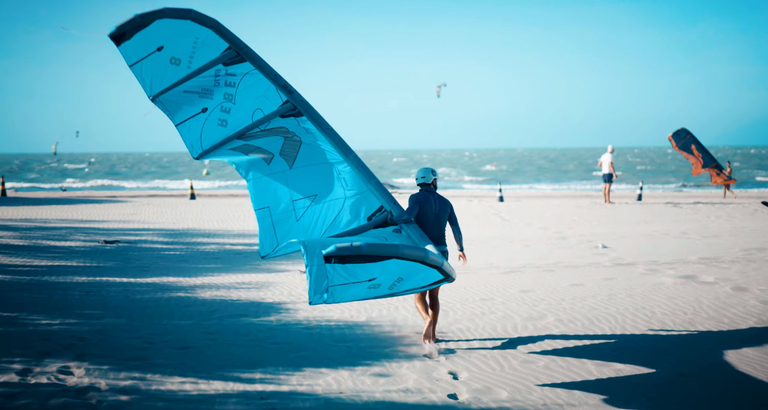 Rubens walks across the beach and toward the water while holding the Duotone Rebel D/LAB kite under his arm.