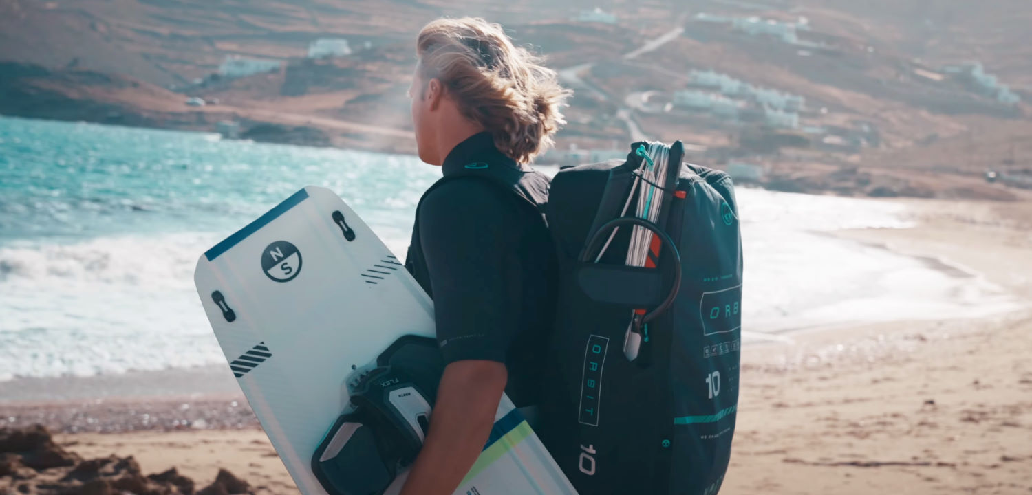 Maxwell Dahl is ready to hit the water. How about you? Maxwell Dahl stands on the beach with a board under his arm and his kite on his back.