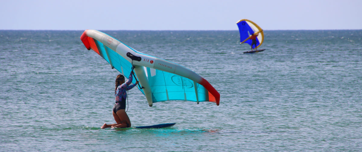 A wing foiler kneels so that the nose of his board rises up just a bit.