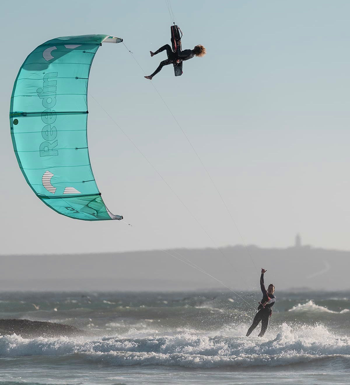 A kiteboarder jumps high while a second kiteboarder rides below him.