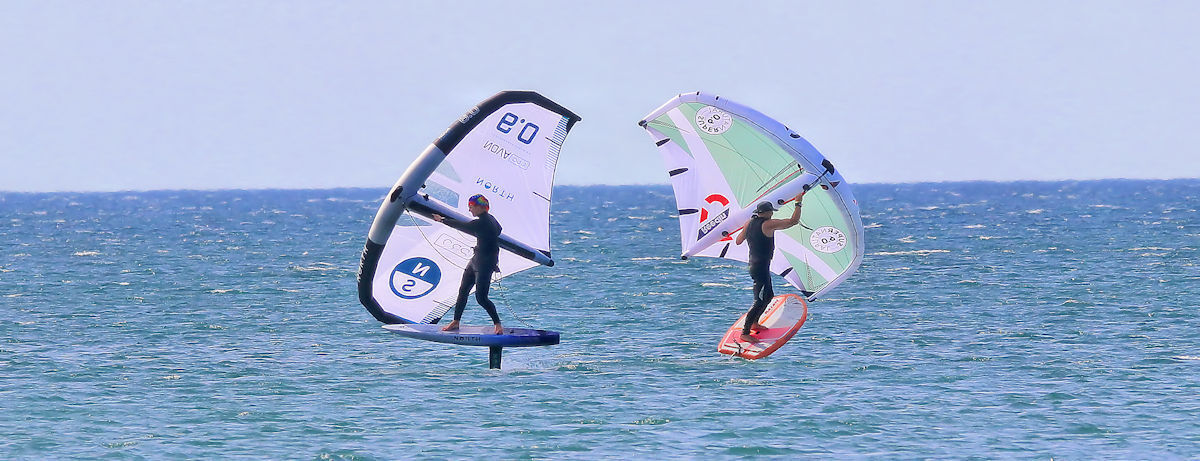 Two wing foilers ride, one with his left foot forward, and the other with his right foot forward.